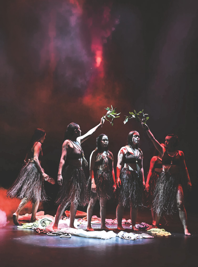 First Nations women dancers in grass skirts holding leaves under red stage light