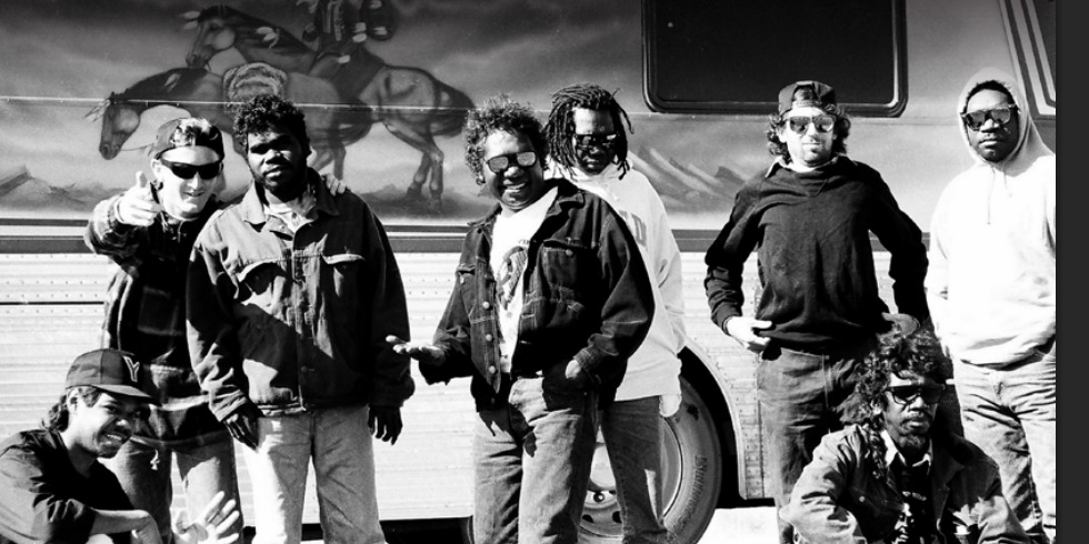 The members of Yothu Yindi standing together in front of their tour bus, black-and-white photograph